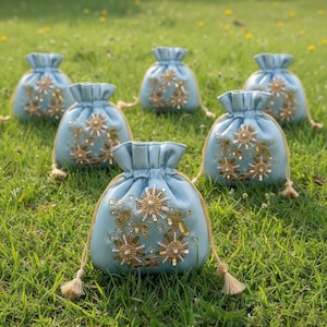Puede incluir: Seis pequeñas bolsas de cordón azul claro con bordados florales dorados y perlados. Cada bolsa tiene una parte superior fruncida y un cordón con borla. Están dispuestas sobre hierba verde.