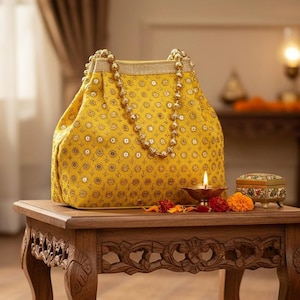 Könnte beinhalten: Eine senfgelbe Handtasche mit Perlenhenkel und goldenen Verzierungen. Die Tasche steht auf einem Holztisch mit Schnitzereien, neben einer kleinen dekorativen Box und einer brennenden Kerze.