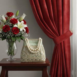 May include: A light green handbag featuring intricate white lace embroidery, small clear beads, and two handles made of large white faux pearls. The bag sits on a wooden surface next to a glass vase filled with red roses and white lilies.