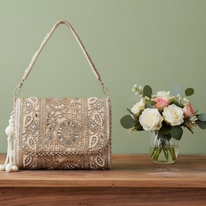 Puede incluir: Bolso de mano beige con correa y borla de cuentas. El bolso está profusamente adornado con perlas y pedrería, con intrincados diseños florales y geométricos. Un pequeño ramo de rosas blancas y rosas está cerca.