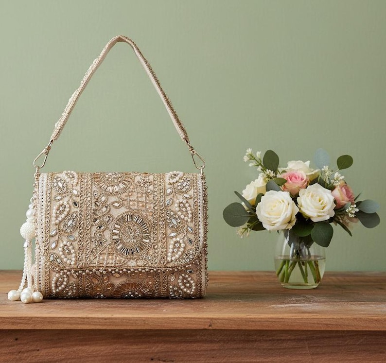 May include: A beige clutch bag with a beaded strap and tassel, adorned with pearls and rhinestones. The bag displays detailed floral and geometric designs. A small vase of white and pink roses is placed beside the bag.