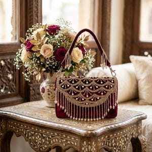 May include: A deep burgundy velvet handbag featuring an intricate gold embroidered scalloped flap with hanging pearl-like beads. The bag includes a matching velvet handle attached with silver-toned metal hardware clips.
