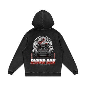 May include: Black hooded sweatshirt with a graphic design featuring a red and white dragon above a black sports car. The text "RISING SUN" is displayed in red and white, with additional Japanese text.