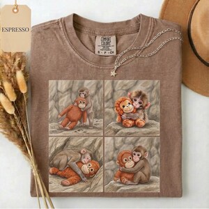 May include: Espresso-colored t-shirt with a four-panel graphic of hugging monkeys. The shirt has a crew neck and short sleeves. A silver necklace with a star pendant is draped on the shirt. The tag reads "Comfort Colors".