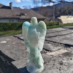 Peut inclure: Une figurine d'ange sculptée dans des tons de vert pâle et de blanc. L'ange a des ailes déployées et se tient sur une surface en bois. L'arrière-plan montre une maison, des arbres et une chaîne de montagnes sous un ciel bleu.