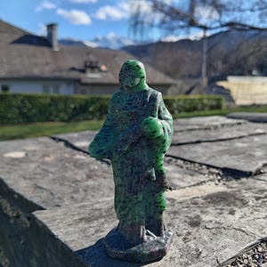 Può includere: Una piccola statua verde di una figura in abito, forse un'icona religiosa, è posta su una superficie di pietra. La statua ha un aspetto strutturato con tonalità verde scuro e rossastre. Lo sfondo presenta una casa, alberi e montagne sotto un cielo luminoso.