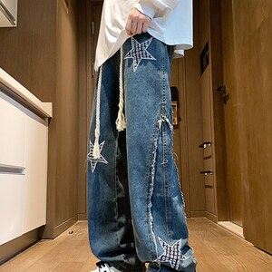 Op de afbeelding: Blauwe spijkerbroek met stervormige patches en een touwkoord. De jeans heeft een relaxte pasvorm en distressed details langs de zijnaden. De sterpatches zijn wit met een zwart patroon.