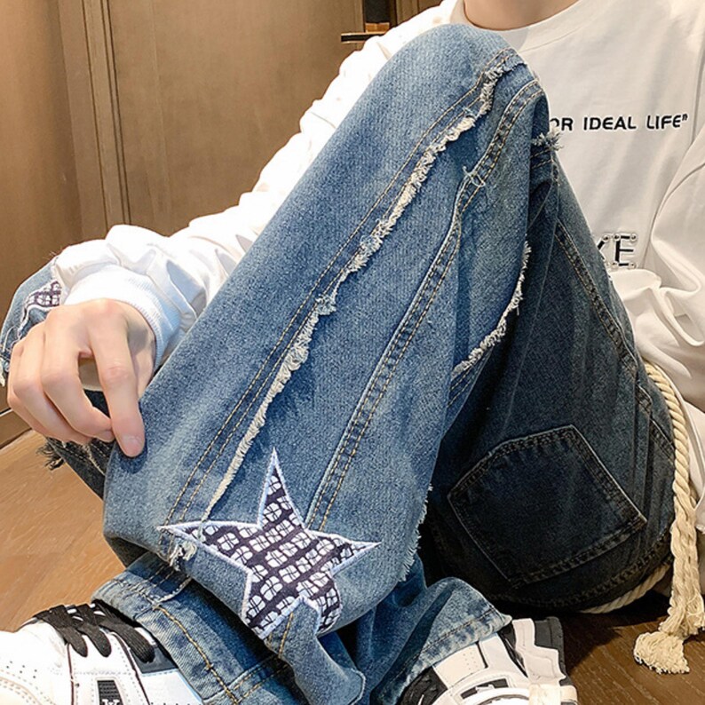 Op de afbeelding: Blauwe spijkerbroek met een sterpatch op de knie en distressed details. De jeans heeft een relaxte pasvorm en een wit trekkoord. De persoon draagt een wit shirt met lange mouwen met de tekst "OR IDEAL LIFE".