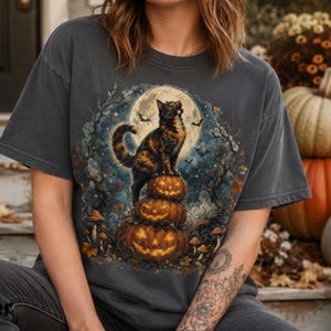 Puede incluir: Una camiseta de manga corta en color gris carbón con un estampado gráfico de un gato carey posado sobre una pila de tres calabazas iluminadas, con un fondo de luna llena, árboles oscuros y murciélagos volando en el cielo nocturno.