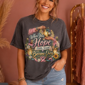 May include: A charcoal gray short-sleeve t-shirt featuring a graphic print of two hands holding a glowing lantern surrounded by flowers and ribbons. The text reads, When Your Hope Feels Tired, Borrow Ours, with pink and yellow floral accents.