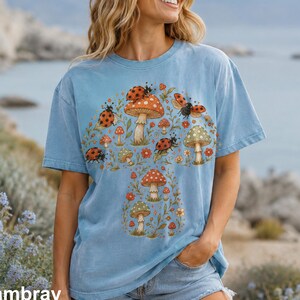 Könnte beinhalten: Ein hellblaues kurzärmeliges T-Shirt mit einem zentralen Grafikdruck aus verschiedenen Pilzen und Marienkäfern. Das Design zeigt mehrere rote und grüne Pilze, umgeben von kleinen orangefarbenen Blumen, grünen Blättern und rot gepunkteten Marienkäfern.
