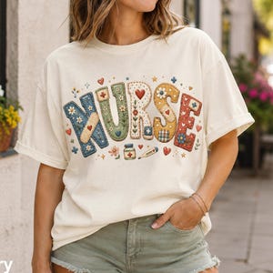 Può includere: Una t-shirt a maniche corte color avorio con la parola NURSE in grandi lettere colorate e decorate. Il design include illustrazioni a tema medico come un cerotto, un flacone di pillole e un termometro, circondati da piccoli cuori e fiori.