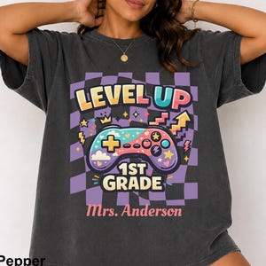 Op de afbeelding: Een antracietgrijs t-shirt met korte mouwen en een grafische print op een paars en zwart geblokte achtergrond. Het ontwerp bevat een kleurrijke gamecontroller, sterren en de tekst "LEVEL UP 1ST GRADE" en "Mrs. Anderson".