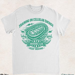 Puede incluir: Una camiseta blanca de manga corta con un estampado gráfico verde de una mitocondria. El texto dice: TRAINING ON CELLULAR ENERGY, ATP ENDURANCE CLUB, KEEP MOVING, 5K y 262. El diseño incluye rayos y alas como elementos gráficos.