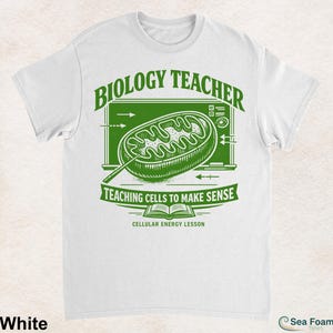 May include: A white t-shirt featuring a green graphic of a mitochondrion on a chalkboard background. The text reads BIOLOGY TEACHER, TEACHING CELLS TO MAKE SENSE, and CELLULAR ENERGY LESSON, accompanied by an open book icon and scientific arrows.