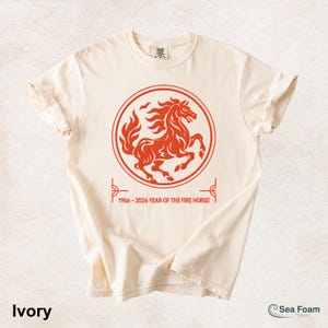 May include: An ivory short-sleeve t-shirt featuring a red circular graphic of a rearing horse. Below the circle, the text reads 1966 - 2026 YEAR OF THE FIRE HORSE. The design is centered on the chest of the shirt, which has a crew neckline.