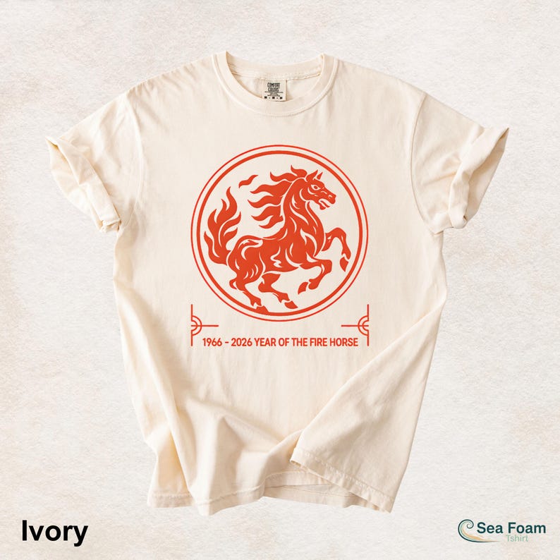 May include: An ivory short-sleeve t-shirt featuring a red circular graphic of a rearing horse. Below the circle, the text reads 1966 - 2026 YEAR OF THE FIRE HORSE. The design is centered on the chest of the shirt, which has a crew neckline.