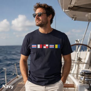Puede incluir: Una camiseta azul marino de manga corta con una fila horizontal de cinco banderas náuticas de señales de colores en el pecho. Las banderas muestran varios patrones geométricos en rojo, blanco, azul y amarillo.
