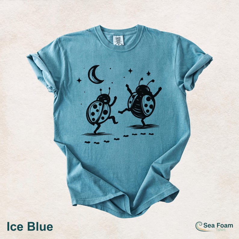 May include: Ice blue t-shirt featuring a black graphic of two dancing ladybugs under a crescent moon and stars. The shirt has a crew neck and short sleeves. The text "Ice Blue" is at the bottom left.
