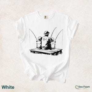 May include: White t-shirt with a black graphic of a person and two children fishing. The person is wearing a hat and the name "Robert" is written on the back. The children's names, "Lucas" and "Noah", are also visible. The shirt also has the word "White" at the bottom.