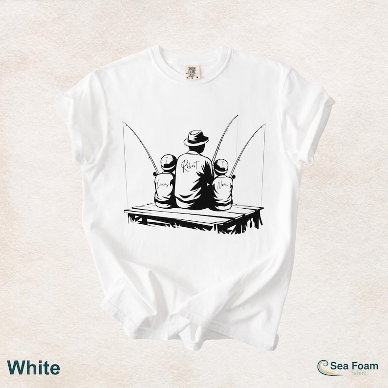 May include: White t-shirt with a black graphic of a person and two children fishing. The person is wearing a hat and the name "Robert" is written on the back. The children's names, "Lucas" and "Noah", are also visible. The shirt also has the word "White" at the bottom.