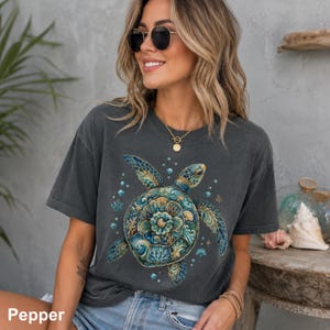 Puede incluir: Una camiseta gris carb&oacute;n de manga corta con un gr&aacute;fico detallado de una tortuga marina con un caparaz&oacute;n ornamentado estilo mandala en tonos azul verdoso, azul y dorado, rodeado de peque&ntilde;as burbujas y detalles de coral en el frente.