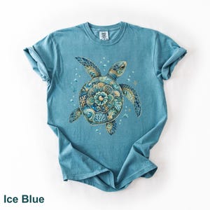 Puede incluir: Una camiseta de manga corta color azul hielo con un gr&aacute;fico central de una tortuga marina cuyo caparaz&oacute;n presenta intrincados patrones florales y de olas en azul, turquesa y dorado, rodeada de peque&ntilde;as burbujas y detalles de coral.