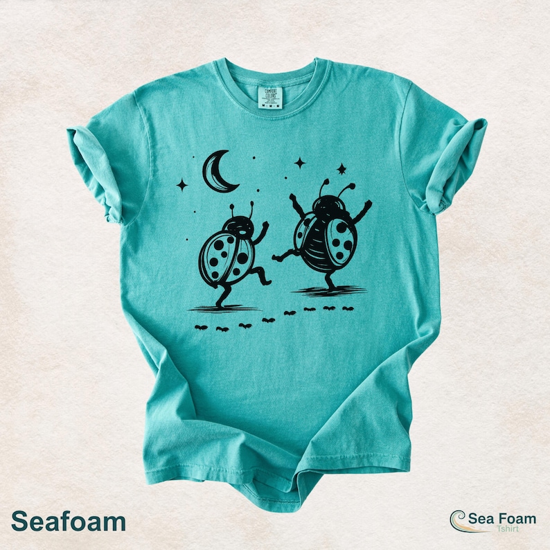 May include: Seafoam green t-shirt with a black graphic of two dancing ladybugs under a crescent moon and stars. The shirt has the word "Seafoam" printed at the bottom.