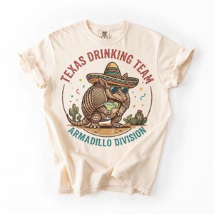 May include: Cream-colored t-shirt featuring a cartoon armadillo wearing sunglasses and a sombrero, holding a cocktail. The shirt has the text "Texas Drinking Team Armadillo Division" in a circular design.