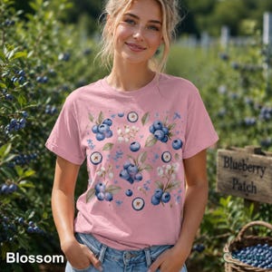 Peut inclure: Un t-shirt rose clair orné d'un motif aquarelle de myrtilles, de feuilles et de fleurs blanches. Le mot "Blossom" est imprimé en bas à gauche. Le t-shirt est à manches courtes et a un col rond.