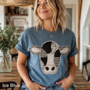 May include: An ice blue t-shirt with a cow face design. The cow face is made of black and white patches, including gingham and floral patterns. The shirt has rolled-up sleeves. The text "Sea Foam T-shirt" is visible.