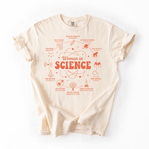 May include: A cream-coloured t-shirt with an orange graphic. The design includes the words "Women in Science" and illustrations of scientists and scientific concepts. The t-shirt is made of a soft material.