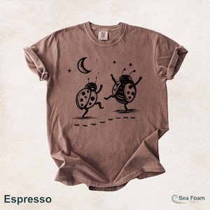 May include: Espresso-colored t-shirt featuring a whimsical design of two dancing ladybugs under a crescent moon and stars. The ladybugs have polka dots and are depicted with arms raised. The shirt has rolled-up sleeves and the text "Espresso".