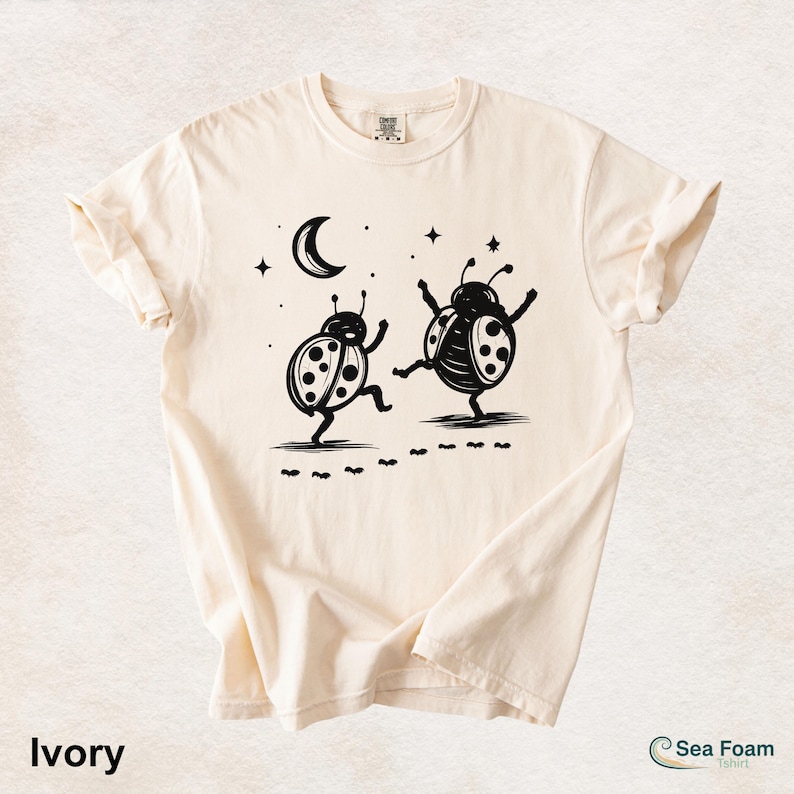 May include: Ivory-colored t-shirt featuring a black illustration of two dancing ladybugs under a crescent moon and stars. The shirt has rolled-up sleeves and the text "Ivory" at the bottom left.