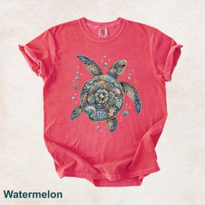 Puede incluir: Una camiseta de manga corta color sand&iacute;a con un gr&aacute;fico central de una tortuga marina. El caparaz&oacute;n de la tortuga muestra intrincados patrones florales y de olas en azul, dorado y turquesa. Peque&ntilde;as burbujas y formas de coral rodean a la tortuga.