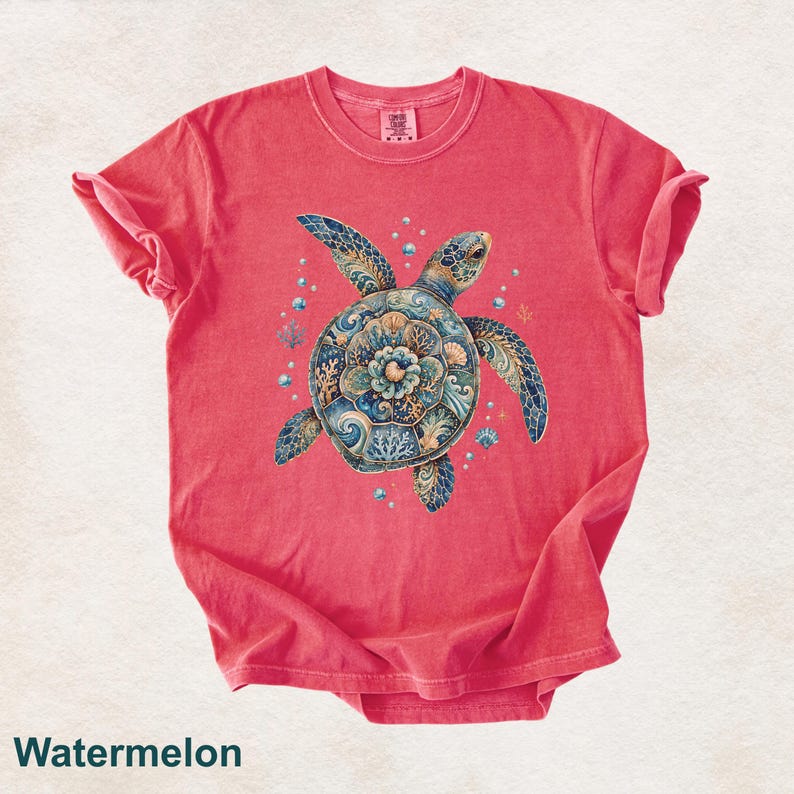 Puede incluir: Una camiseta de manga corta color sand&iacute;a con un gr&aacute;fico central de una tortuga marina. El caparaz&oacute;n de la tortuga muestra intrincados patrones florales y de olas en azul, dorado y turquesa. Peque&ntilde;as burbujas y formas de coral rodean a la tortuga.