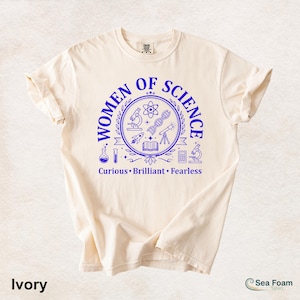 以下が含まれることがあります： アイボリー色のTシャツ。青い円形のグラフィックには「WOMEN OF SCIENCE」の文字と科学関連のイラストが描かれています。「Curious, Brilliant, Fearless」の文字が下にあります。Sea Foam製です。
