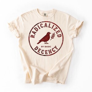 Può includere: T-shirt color crema con grafica circolare bordeaux. La grafica presenta la silhouette di un uccello, un rametto e le parole "RADICALIZED" e "DECENCY" che circondano l'uccello, con "BY BASIC" sotto. La maglietta ha le maniche arrotolate.