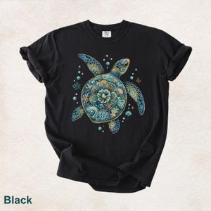 Puede incluir: Una camiseta negra con un gr&aacute;fico central de una tortuga marina con un caparaz&oacute;n compuesto por intrincados patrones florales y de coral en tonos azul, verde azulado y dorado. El dise&ntilde;o incluye peque&ntilde;as burbujas, estrellas de mar y conchas.