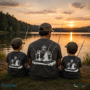 May include: Three people wearing gray t-shirts with a fishing-themed graphic, sitting by a lake at sunset. The graphic features a fisherman with two fishing poles and the word "Reel" on the shirt. The scene is peaceful.