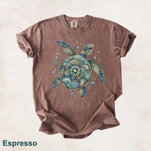 Puede incluir: Una camiseta de manga corta color marr&oacute;n espresso con un gr&aacute;fico central de una tortuga marina. El caparaz&oacute;n de la tortuga est&aacute; decorado con intrincados patrones florales y oce&aacute;nicos en azul, turquesa y dorado, rodeado de burbujas y corales.