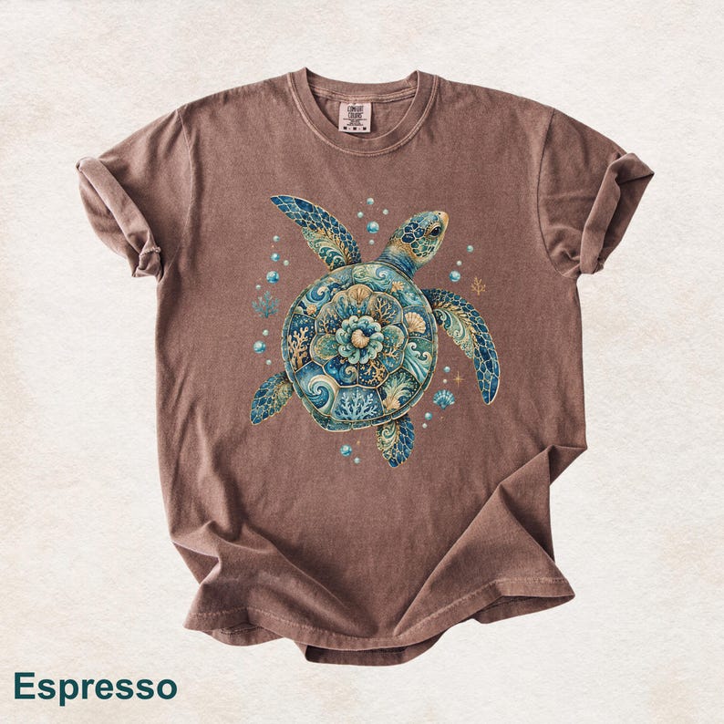 Puede incluir: Una camiseta de manga corta color marr&oacute;n espresso con un gr&aacute;fico central de una tortuga marina. El caparaz&oacute;n de la tortuga est&aacute; decorado con intrincados patrones florales y oce&aacute;nicos en azul, turquesa y dorado, rodeado de burbujas y corales.
