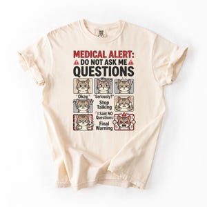 May include: Cream-colored t-shirt with the text "MEDICAL ALERT: DO NOT ASK ME A QUESTIONS." Below the text are six panels with cat illustrations expressing different emotions. The shirt has a crew neck and short sleeves.