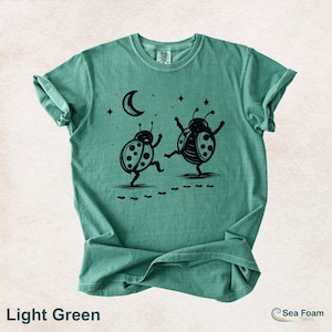May include: Light green t-shirt with a black graphic of two dancing ladybugs under a crescent moon and stars. The shirt has rolled-up sleeves and the text "Light Green" at the bottom.
