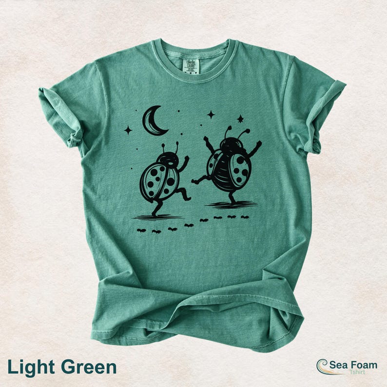 May include: Light green t-shirt with a black graphic of two dancing ladybugs under a crescent moon and stars. The shirt has rolled-up sleeves and the text "Light Green" at the bottom.