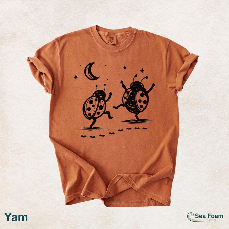 May include: A burnt orange t-shirt featuring a black graphic of two dancing ladybugs under a crescent moon and stars. The shirt has rolled-up sleeves and the word "Yam" is printed at the bottom left. The Sea Foam t-shirt logo is at the bottom right.