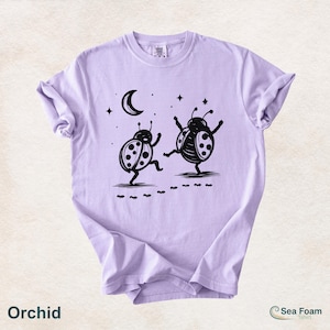 May include: Orchid-colored t-shirt featuring a whimsical black and white illustration of two dancing ladybugs under a crescent moon and stars. The shirt has a crew neck and short sleeves. The text "Orchid" is at the bottom.