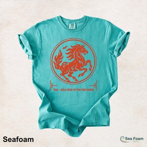 May include: A seafoam green short-sleeve t-shirt featuring a circular orange graphic of a rearing horse. Below the circle, the text reads 1966 - 2026 YEAR OF THE FIRE HORSE. The shirt has a crew neckline and a standard fit with a folded sleeve detail.