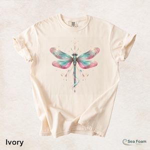May include: Ivory-colored t-shirt featuring a watercolor dragonfly design. The dragonfly has teal and pink wings with gold accents. The shirt has a crew neck and short sleeves. The text "Ivory" is at the bottom left.