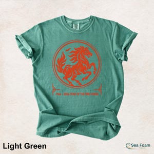May include: A light green short-sleeve t-shirt featuring a circular orange graphic of a rearing horse. Below the circle, the text reads 1966 - 2026 YEAR OF THE FIRE HORSE. The shirt has a crew neckline and a standard fit with a soft, washed texture.